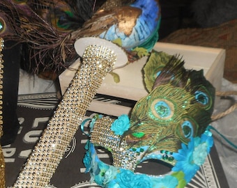 May include: A blue and gold peacock feather mask with turquoise and green accents. The mask is decorated with blue and green flowers and has a peacock feather design.