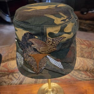 May include: Camouflage-patterned cap with decorative embellishments. The cap features a brown and gold eagle emblem, along with silver and brown decorative accents. The cap is displayed on a gold stand.