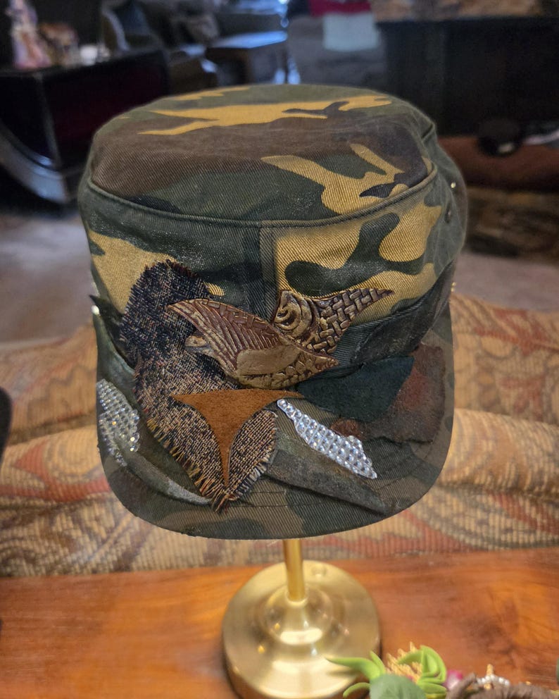 May include: Camouflage-patterned cap with decorative embellishments. The cap features a brown and gold eagle emblem, along with silver and brown decorative accents. The cap is displayed on a gold stand.