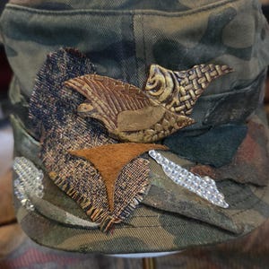 May include: Camouflage-patterned cap with decorative patches and embellishments. Features brown leather and textured fabric accents, along with silver rhinestone details. The cap's design includes a bird-like emblem and a geometric shape.