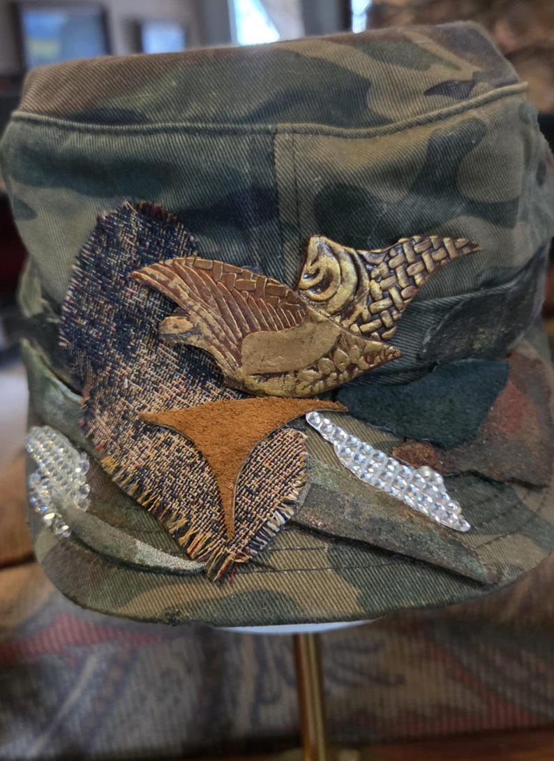 May include: Camouflage-patterned cap with decorative patches and embellishments. Features brown leather and textured fabric accents, along with silver rhinestone details. The cap's design includes a bird-like emblem and a geometric shape.