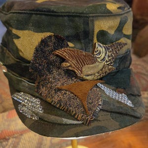 May include: Camouflage print cap with decorative embellishments. Features a brown, textured bird-like applique, a patch of dark fabric, and a line of clear, sparkling rhinestones. The cap is set on a gold-colored stand.
