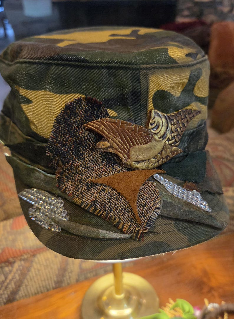 May include: Camouflage print cap with decorative embellishments. Features a brown, textured bird-like applique, a patch of dark fabric, and a line of clear, sparkling rhinestones. The cap is set on a gold-colored stand.