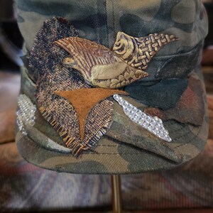 May include: Camouflage-patterned cap with decorative embellishments. Features a brown leather-like bird design, fabric patches, and silver rhinestone accents. The cap is displayed on a gold-colored stand.