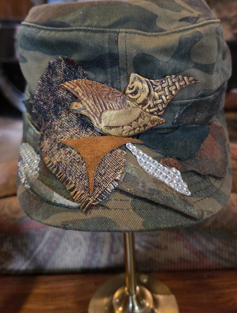 May include: Camouflage-patterned cap with decorative embellishments. Features a brown leather-like bird design, fabric patches, and silver rhinestone accents. The cap is displayed on a gold-colored stand.