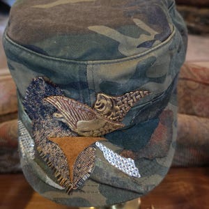 May include: Camouflage-patterned military-style cap with decorative embellishments. The cap features a brown and gold-colored emblem, along with patches of brown, silver, and textured fabric. The cap is displayed on a gold-colored stand.
