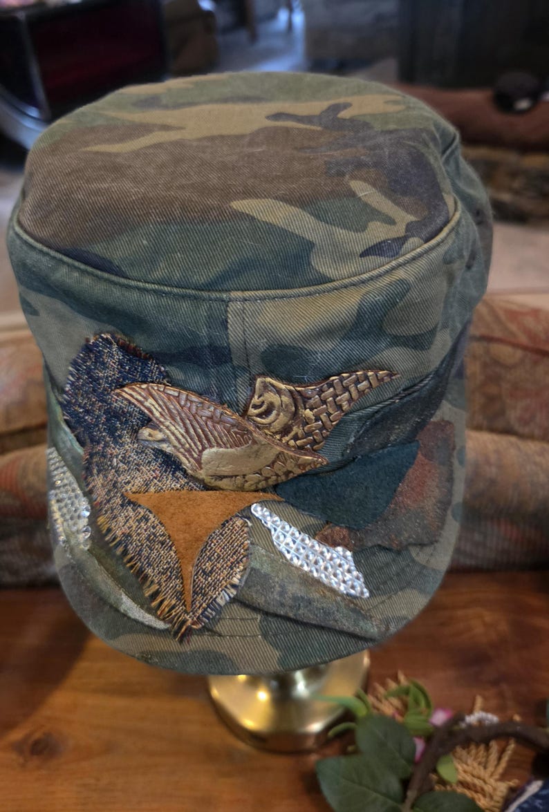 May include: Camouflage-patterned military-style cap with decorative embellishments. The cap features a brown and gold-colored emblem, along with patches of brown, silver, and textured fabric. The cap is displayed on a gold-colored stand.