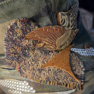 May include: Camouflage-patterned hat with decorative patches and embellishments. Features a textured, multi-colored fabric patch, a brown leather-like star, and a carved wooden accent. The hat is adorned with small, sparkling rhinestones.