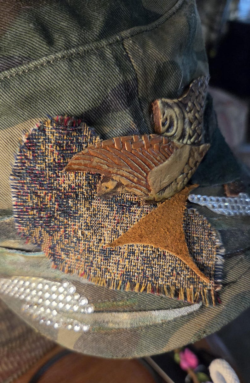 May include: Camouflage-patterned hat with decorative patches and embellishments. Features a textured, multi-colored fabric patch, a brown leather-like star, and a carved wooden accent. The hat is adorned with small, sparkling rhinestones.