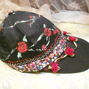 May include: A black baseball cap with a floral design and a colorful, multi-colored ribbon. The cap has a black brim and is decorated with red roses and green leaves.