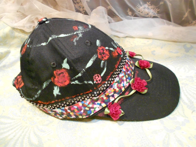 May include: A black baseball cap with a floral design and a colorful, multi-colored ribbon. The cap has a black brim and is decorated with red roses and green leaves.
