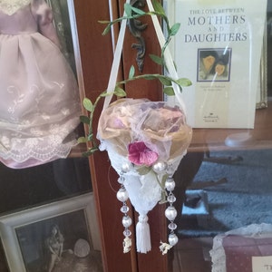 May include: A decorative hanging basket with a white lace base, adorned with pearls, crystals, and a white tassel. The basket is decorated with pink and beige flowers and greenery. A book titled "The Love Between Mothers and Daughters" is in the background.