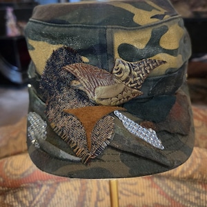 May include: Camouflage military-style cap with decorative embellishments. The cap features a mix of green, brown, and tan fabric. Embellishments include a brown leather-like bird, fabric patches, and silver-colored rhinestones.