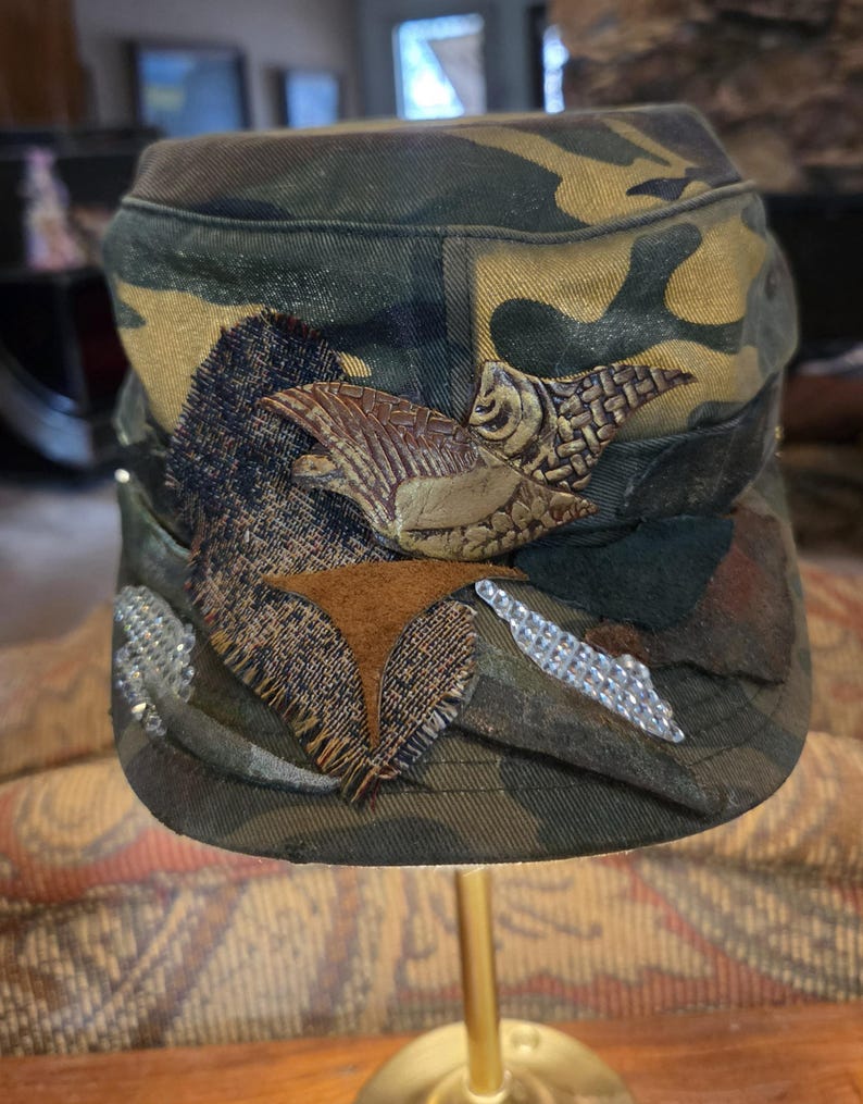 May include: Camouflage military-style cap with decorative embellishments. The cap features a mix of green, brown, and tan fabric. Embellishments include a brown leather-like bird, fabric patches, and silver-colored rhinestones.