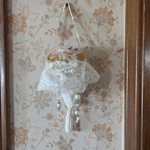 May include: A decorative hanging ornament with a white lace skirt, pearl accents, and a tassel. The ornament is suspended from a white ribbon against a floral wallpaper background.