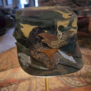 May include: Camouflage-patterned cap with decorative patches and embellishments. The cap features brown, tan, and green hues, with textured fabric patches and metallic accents. The design includes a fish-shaped emblem and a star-shaped patch.