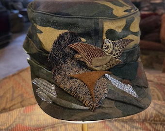 May include: Camouflage-patterned cap with decorative patches and embellishments. The cap features brown, tan, and green hues, with textured fabric patches and metallic accents. The design includes a fish-shaped emblem and a star-shaped patch.