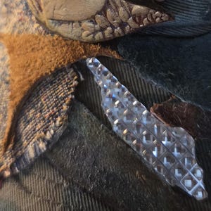 May include: Close-up of a hat with decorative elements. The hat features a textured, dark green fabric, with brown and gold embellishments. A clear, diamond-patterned accent is also visible.