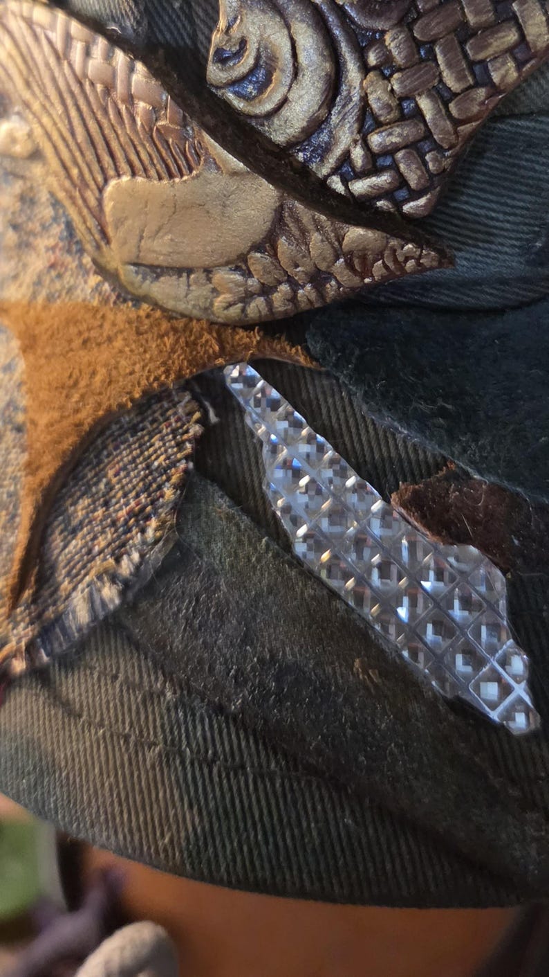 May include: Close-up of a hat with decorative elements. The hat features a textured, dark green fabric, with brown and gold embellishments. A clear, diamond-patterned accent is also visible.