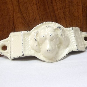 May include: White cast iron drawer pull with a dog's head in the center. The pull has a rectangular shape with rounded edges and a textured surface.