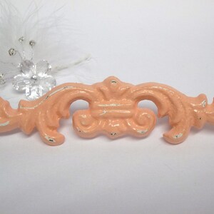May include: A peach-colored metal decorative accent with a distressed finish. The accent has a scroll-like design with a central flourish.