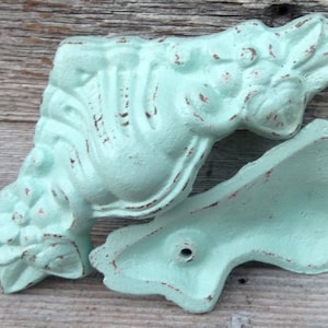 May include: Two antique-style, light blue, cast iron, decorative brackets with a distressed finish. The brackets have a floral design and one has a hole in the center.