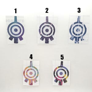 May include: Five colorful vinyl stickers featuring a stylized, geometric design with a central circular element and a rectangular top. The designs vary in color, including blue, purple, and a rainbow pattern. The stickers are on a clear backing.