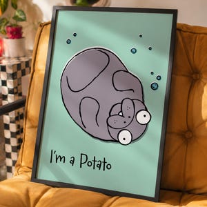 Op de afbeelding: Ingelijste kunstprint met een grillige illustratie van een grijze aardappel met grote ogen en de tekst "I'm a Potato" op een lichtgroene achtergrond. Het kunstwerk is ingelijst in een zwarte lijst.