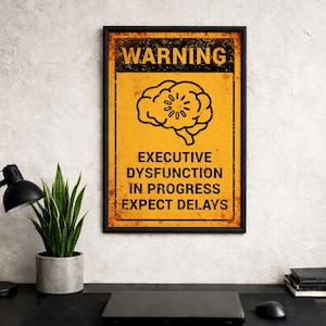 May include: A framed poster with a yellow background and black text that reads "WARNING EXECUTIVE DYSFUNCTION IN PROGRESS EXPECT DELAYS." The poster features a brain graphic and a distressed, vintage style.