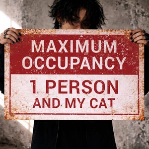 Op de afbeelding: Een rechthoekig metalen bord met een verweerde, roestige uitstraling. Het bord heeft een rood bovenstuk met de witte woorden "MAXIMUM OCCUPANCY". Daaronder staat in een wit gedeelte "1 PERSON AND MY CAT".