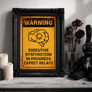 May include: A framed sign with the text "WARNING" and "EXECUTIVE DYSFUNCTION IN PROGRESS EXPECT DELAYS" on a yellow background. The frame is black and ornate. A black plush bunny and candles are nearby.