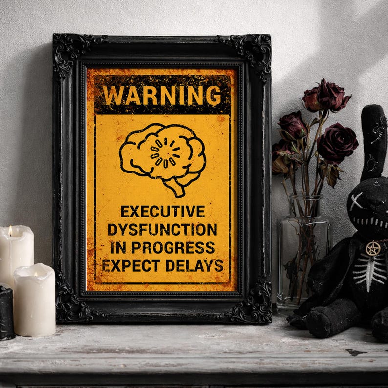 May include: A framed sign with the text "WARNING" and "EXECUTIVE DYSFUNCTION IN PROGRESS EXPECT DELAYS" on a yellow background. The frame is black and ornate. A black plush bunny and candles are nearby.