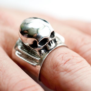 May include: A silver skull ring with a detailed design. The skull is the main feature of the ring, and it is set on a band with a textured surface.