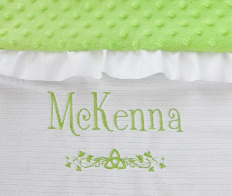 May include: White baby blanket with green embroidery of the name McKenna and a Celtic knot with shamrocks.
