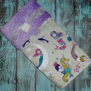 May include: A soft, purple minky baby blanket with a white satin ribbon. The blanket features a whimsical design of mermaids, seahorses, and stars on a light grey background.