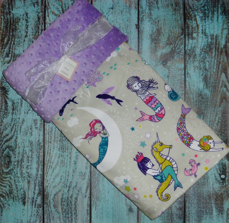 May include: A soft, purple minky baby blanket with a white satin ribbon. The blanket features a whimsical design of mermaids, seahorses, and stars on a light grey background.