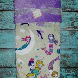 May include: A purple minky baby blanket with a beige fabric side featuring a mermaid theme. The beige side has a pattern of mermaids, seahorses, stars, and seashells. The blanket has a satin ribbon tied around it.
