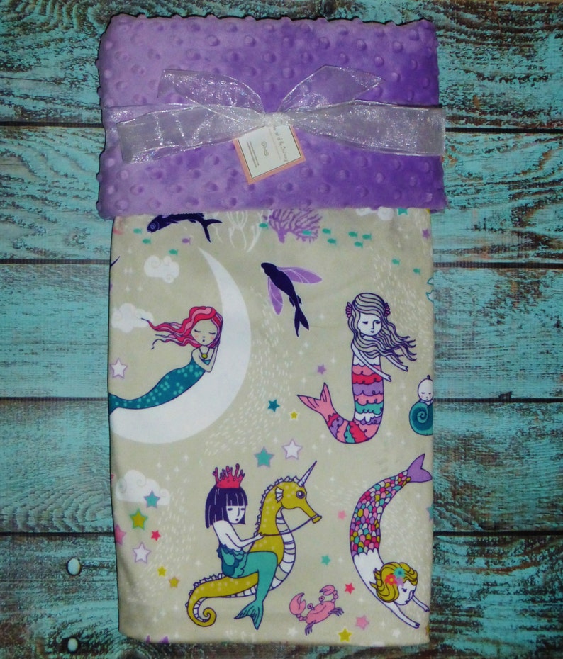 May include: A purple minky baby blanket with a beige fabric side featuring a mermaid theme. The beige side has a pattern of mermaids, seahorses, stars, and seashells. The blanket has a satin ribbon tied around it.