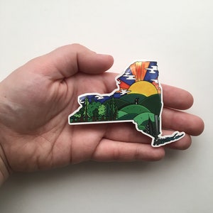 May include: A sticker shaped like the state of New York with a colorful sunrise over a green landscape and a silhouette of a couple in the foreground.
