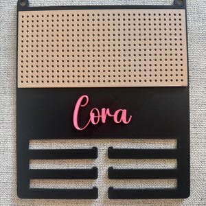 May include: A black wall-mounted display rack featuring a tan pegboard section at the top, the name Cora in pink script lettering in the center, and four horizontal slots at the bottom for hanging items, with two circular mounting holes at the top.