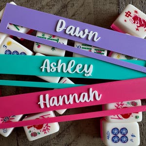 May include: Three rectangular plastic mahjong tile racks in purple, teal, and pink, each featuring a raised white script name: Dawn, Ashley, and Hannah. The racks are layered over a collection of white mahjong tiles with red, green, and blue designs.