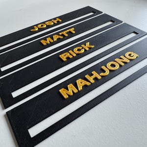 May include: Four black rectangular nameplates with gold lettering. The names "JOSH", "MATT", "RICK", and "MAHJONG" are displayed. Each plate has a slot cut out of the center. The plates are arranged on a white surface.