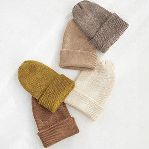 May include: Four knitted beanies in different colours: brown, beige, cream, and mustard yellow. The beanies are folded and arranged on a white surface.