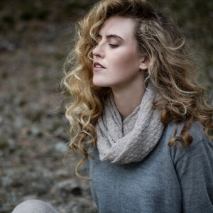May include: A woman with long, curly blonde hair wearing a gray sweater and a light brown scarf. She is looking down with her eyes closed.