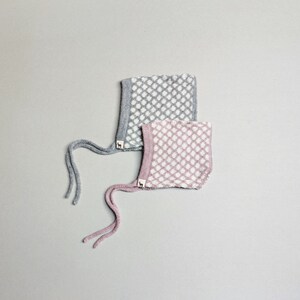 Può includere: Due berretti per neonati in maglia con pois bianchi su sfondo grigio e rosa. I berretti hanno lacci che possono essere legati sotto il mento.