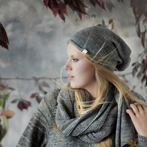 May include: A woman wearing a gray knit sweater and a gray beanie with a scarf. She is standing in front of a gray background with red leaves.