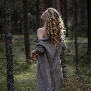 Puede incluir: Una mujer con el pelo largo y rizado rubio se encuentra en un bosque, llevando un c&aacute;rdigan marr&oacute;n. Est&aacute; mirando hacia arriba a los &aacute;rboles.