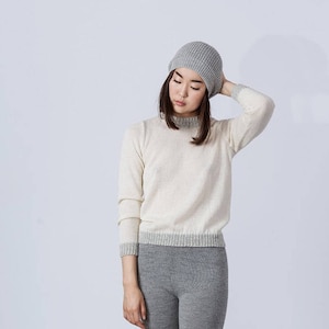 May include: A woman wearing a white sweater with a grey trim and grey beanie. She is wearing grey pants.