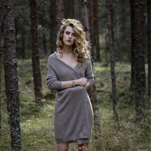 Puede incluir: Una mujer con el pelo largo y rizado rubio se encuentra en un bosque. Lleva un c&aacute;rdigan largo de punto marr&oacute;n y una pulsera en la mu&ntilde;eca. El bosque est&aacute; lleno de &aacute;rboles altos y hierba verde.