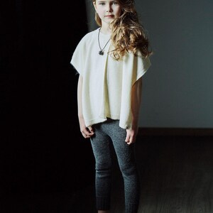 May include: A young girl with long blonde hair wearing a white sweater and grey pants. She is standing in front of a dark background and looking at the camera.